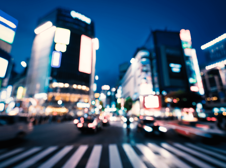 A blurred, vibrant cityscape at night featuring a busy intersection. The scene includes illuminated buildings with large digital billboards and signs, creating a colorful and dynamic atmosphere. The lights from cars and streetlights add to the bustling urban environment, emphasizing the movement and energy of the city.