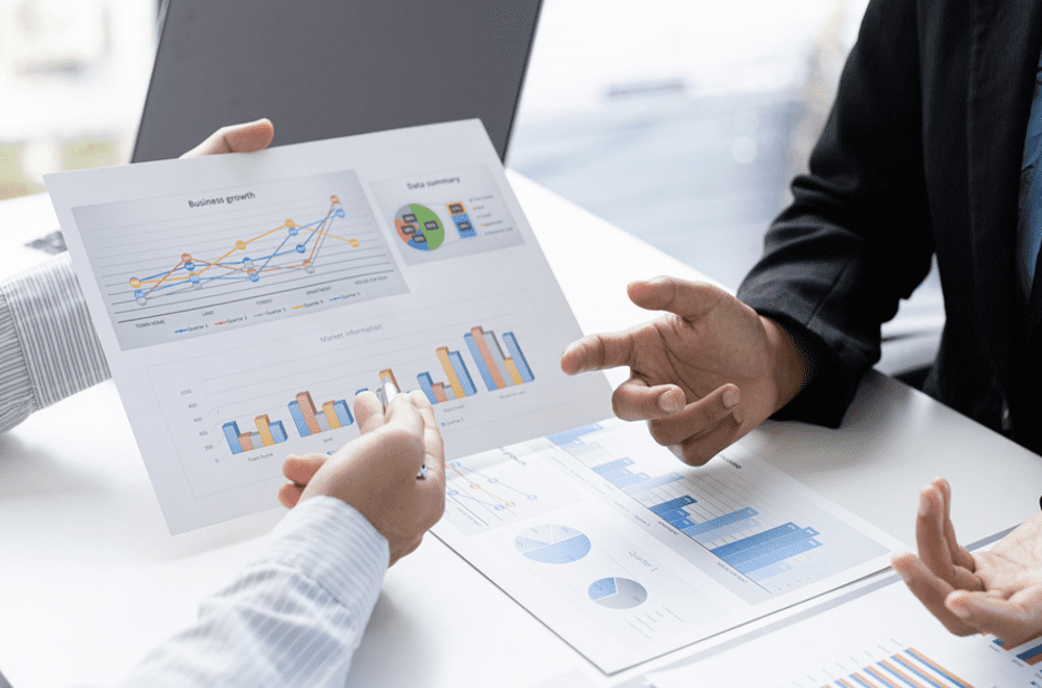 Two business professionals are engaged in a discussion, focusing on a sheet of paper with various charts and graphs. The paper displays line graphs, bar charts, and pie charts, indicating data analysis and business growth. One person is pointing at the charts, while the other is gesturing with their hands, suggesting an active conversation about the data