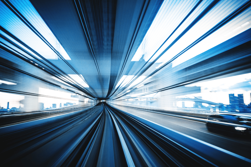 Dynamic, high-speed view of a transportation tunnel or track, creating a sense of rapid motion. The image features converging lines and a blurred effect, suggesting fast movement through an urban environment. Buildings and structures can be seen through the windows, enhanced by the blue and white color tones, contributing to the futuristic and energetic feel of the scene