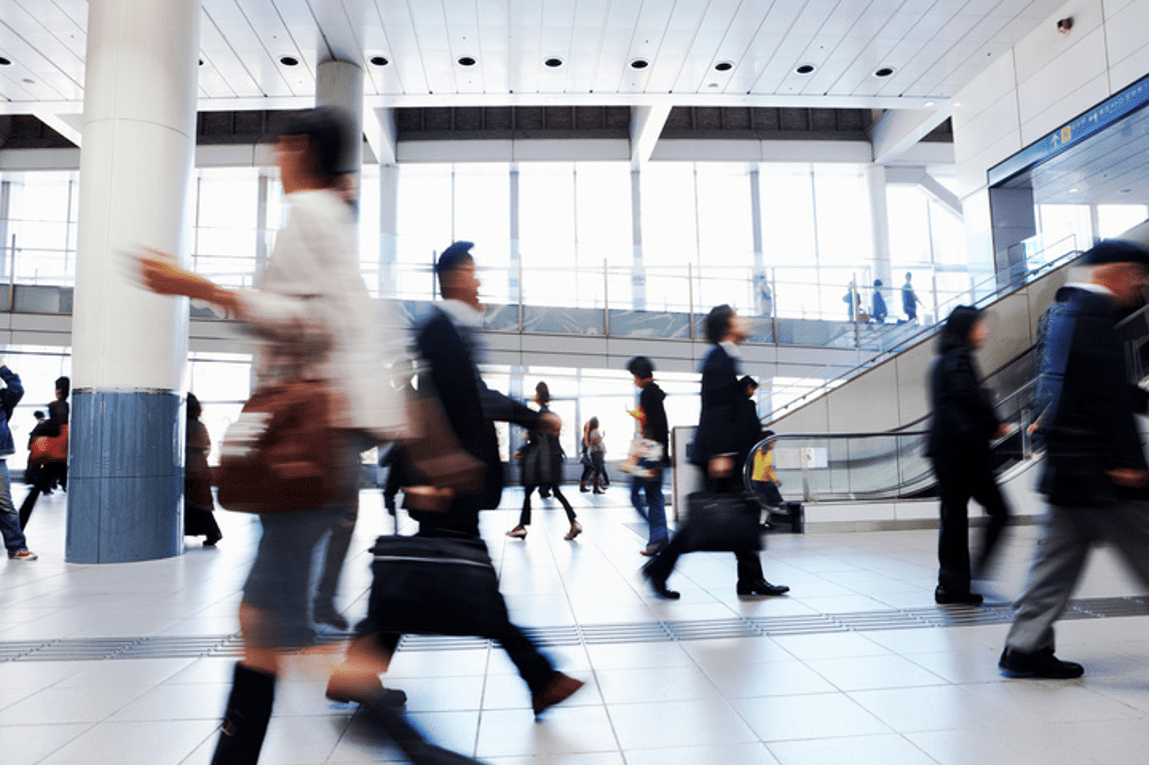 A busy indoor space with people walking in various directions, suggesting a transportation hub or public building. The scene is slightly blurred to emphasize motion, and the architecture features large windows, high ceilings, and pillars. The individuals are dressed in a mix of casual and business attire, carrying bags and briefcases, contributing to the bustling atmosphere