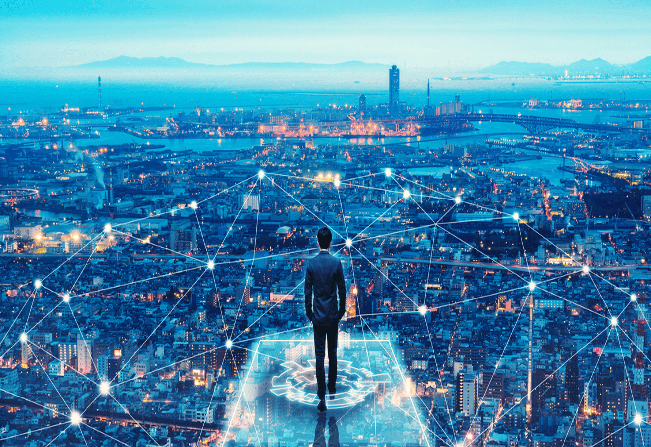 A man in a suit stands on a platform overlooking a vast cityscape at dusk. The city below is illuminated with lights, and a network of interconnected lines and nodes overlay the scene, symbolizing connectivity and data networks. In the background, there are buildings, bridges, and a body of water, with mountains visible on the horizon. The image conveys a sense of futuristic technology and global connection