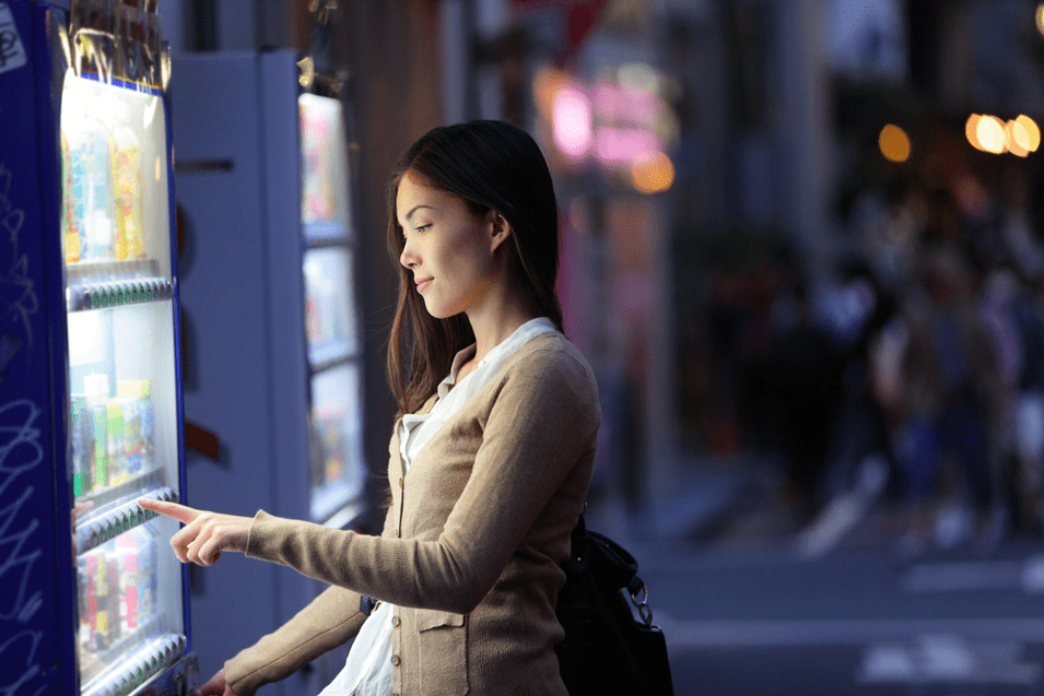 A young woman is standing in front of a vending machine on a city street at dusk. She is dressed in a beige cardigan and white blouse, and is pointing at the machine to make a selection. The background is softly blurred, showing other vending machines and people walking, with city lights creating a warm, urban atmosphere